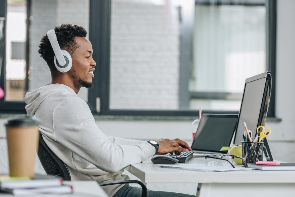 χαμόγελο Αφρικανός Αμερικανός προγραμματιστής στα ακουστικά που κάθονται στο χώρο εργασίας στο γραφείο - Φωτογραφία, εικόνα