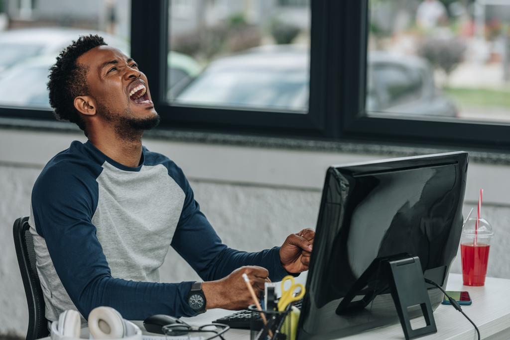сердитый африканский американский программист жестикулирует, сидя на рабочем месте в офисе
 - Фото, изображение