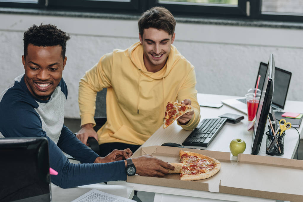 ofiste masalarda otururken pizza yiyen iki neşeli çok kültürlü programcılar - Fotoğraf, Görsel