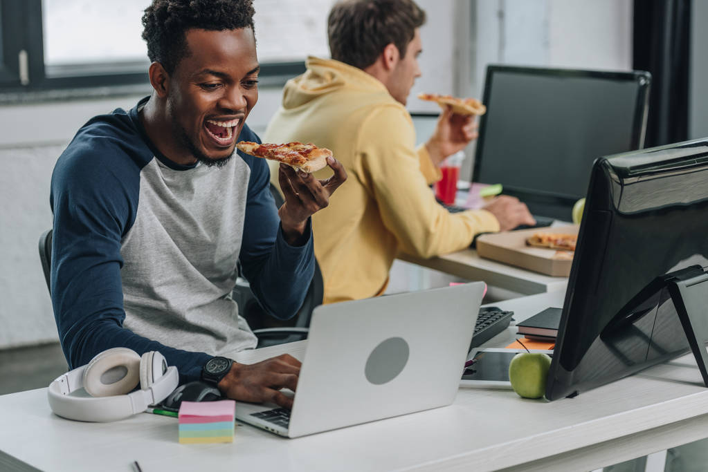 два мультикультурних програмісти їдять піцу, сидячи біля комп'ютерів в офісі
 - Фото, зображення
