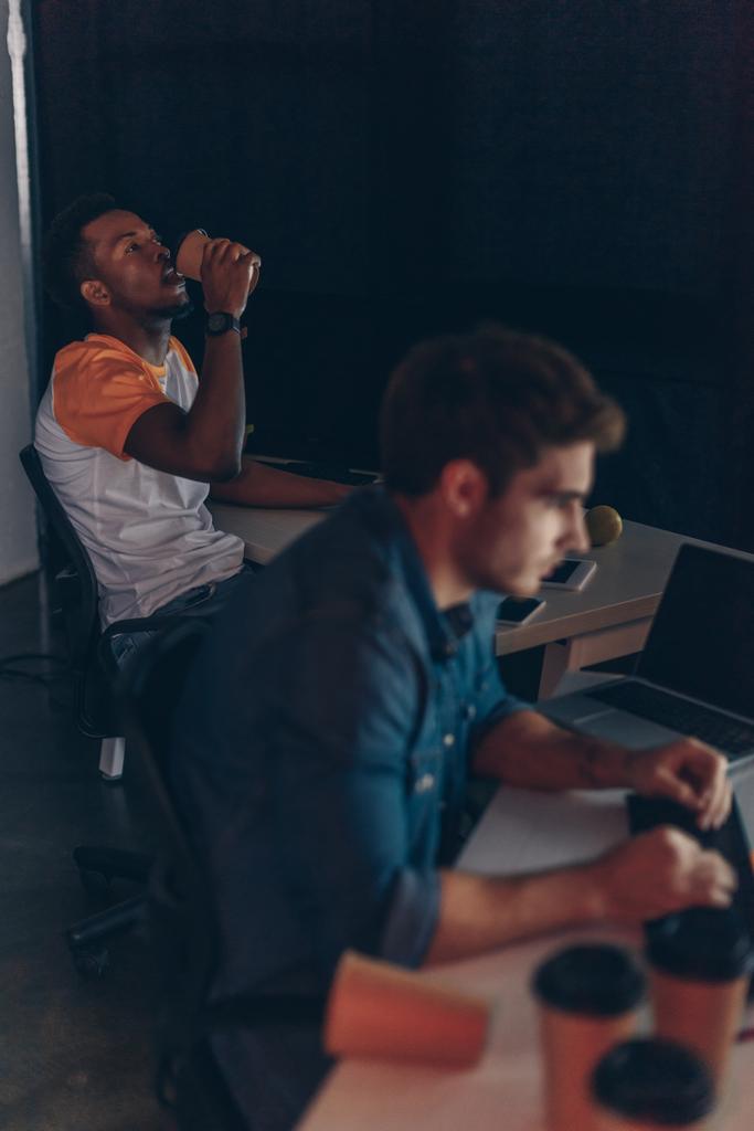 Selektywny fokus African American programista picia kawy, aby przejść podczas pracy w pobliżu wielokulturowego kolegę w nocy w biurze - Zdjęcie, obraz
