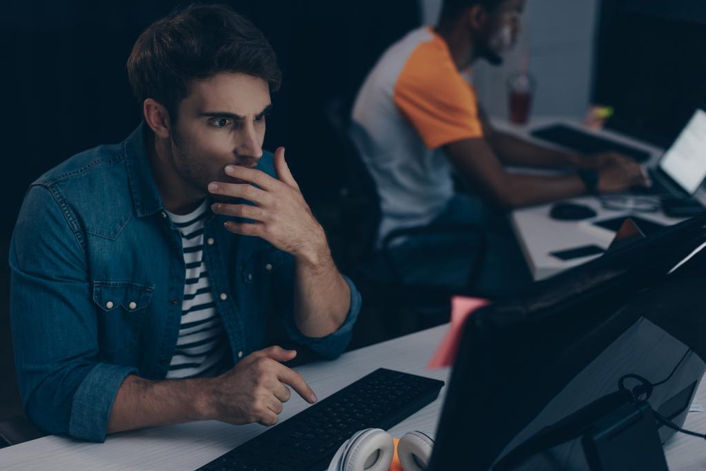 selective focus of shocked programmer working at night in office near multicultural friend - Photo, Image