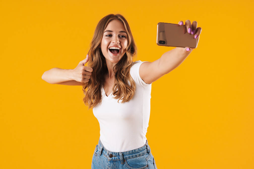 Imagen de primer plano de la alegre mujer caucásica que usa una camiseta básica que muestra el pulgar hacia arriba mientras toma una foto selfie en el teléfono celular
 - Foto, imagen