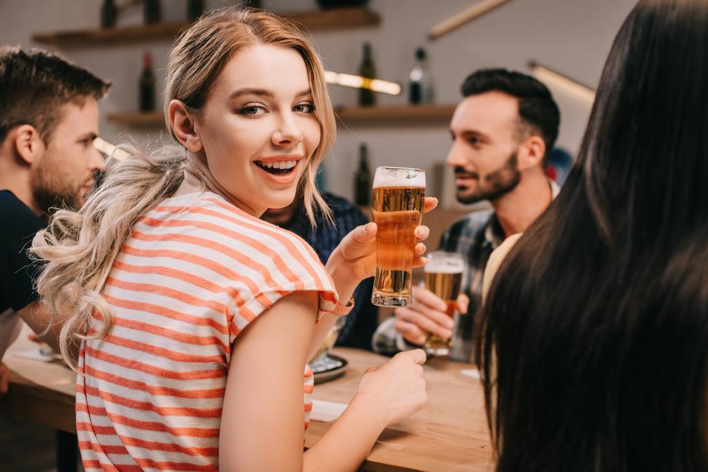 selektywne skupienie radosnej młodej kobiety uśmiechając się przy kamerze, trzymając szklankę lekkiego piwa - Zdjęcie, obraz
