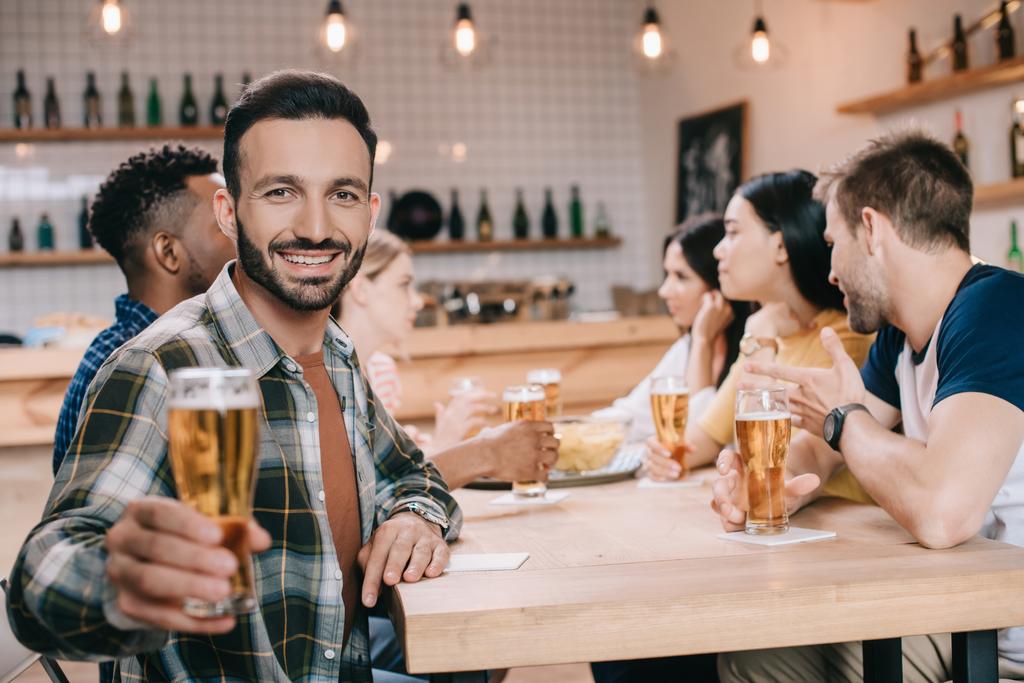 επιλεκτική εστίαση του όμορφου άντρα που χαμογελά στην κάμερα ενώ κρατά ένα ποτήρι μπύρα  - Φωτογραφία, εικόνα