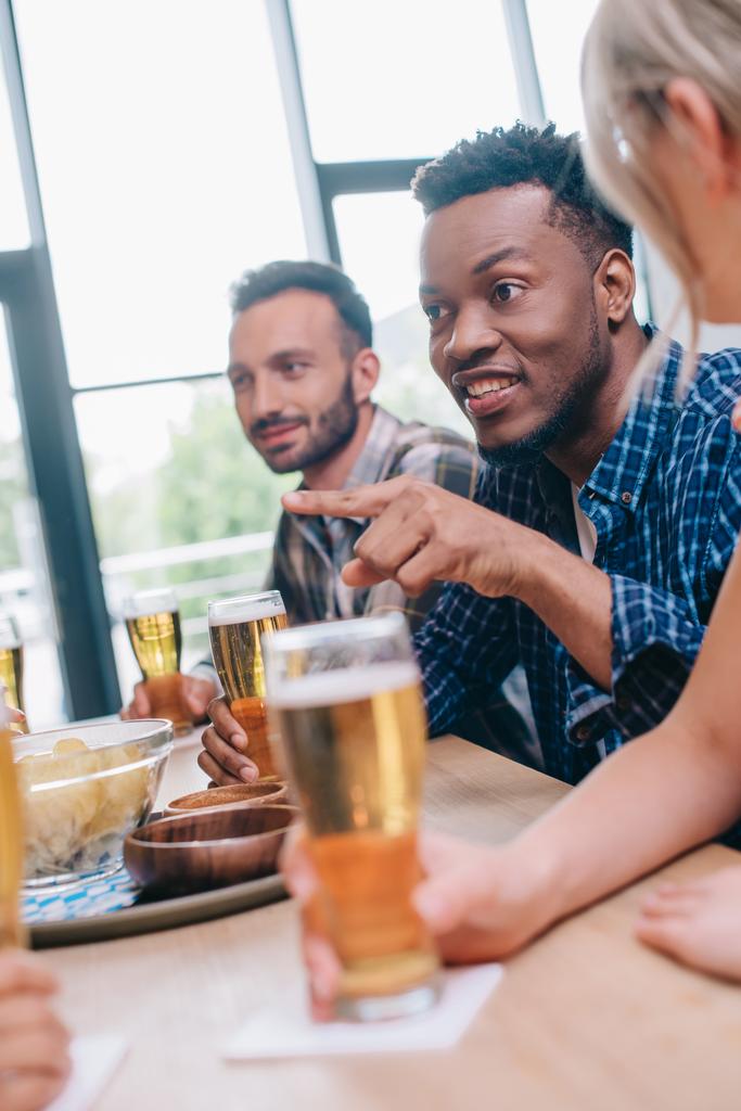 selective focus of cheerful african american man pointing with finger while sitting with multicultural friends in pub - Photo, Image