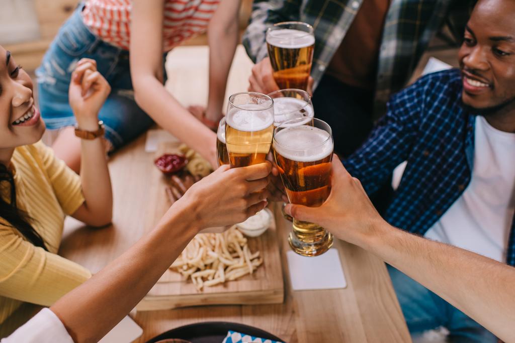 vista recortada de amigos multiculturales tintineando vasos de cerveza ligera en el pub
 - Foto, Imagen