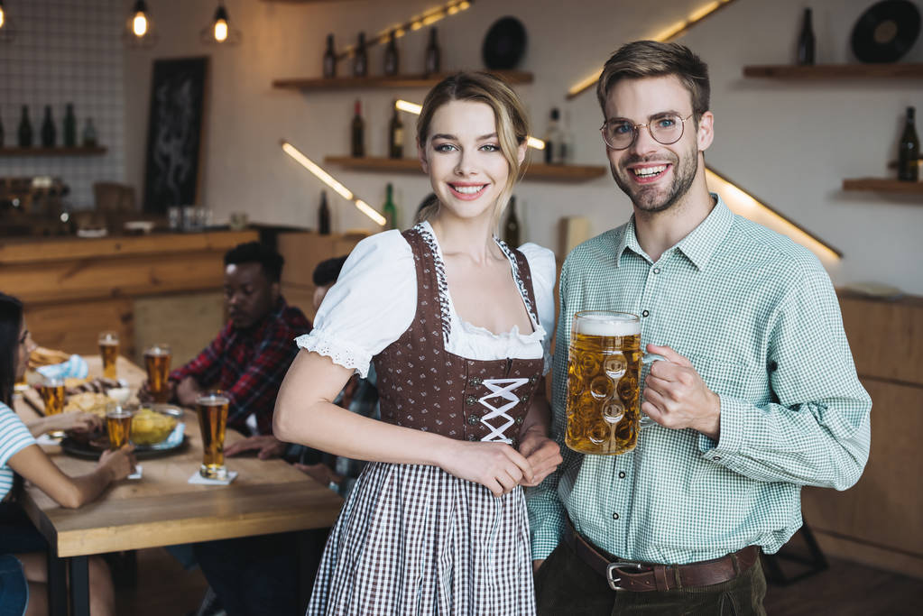 hermosa camarera en traje nacional alemán de pie cerca de hombre joven sosteniendo taza de cerveza ligera y sonriendo a la cámara
 - Foto, Imagen