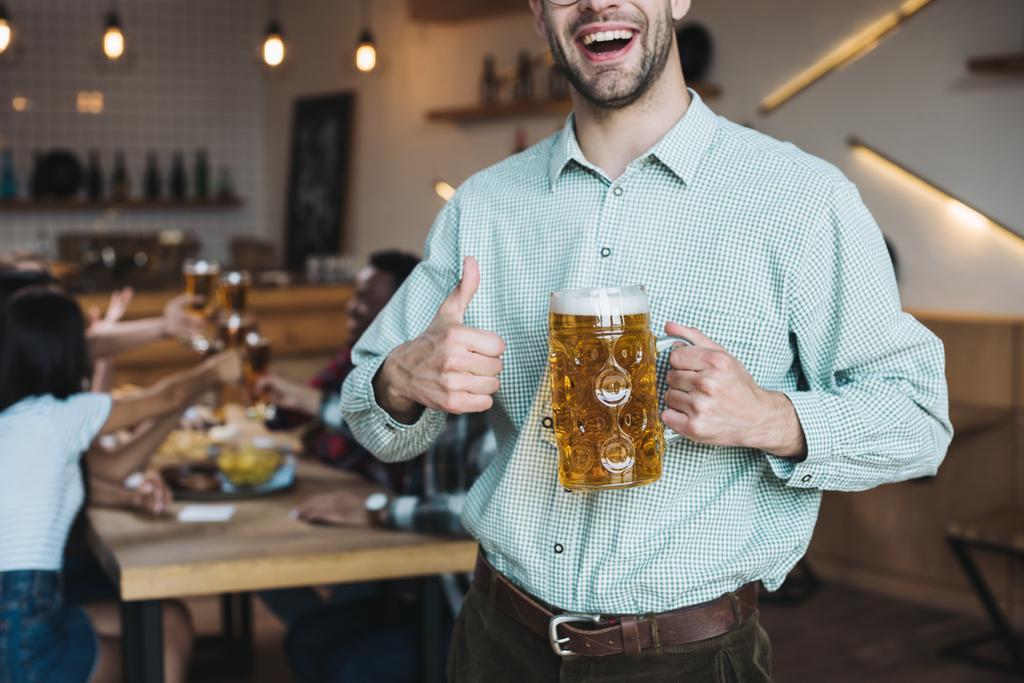 nyírt kilátás mosolygó fiatalember gazdaság bögre lager sör és mutatja hüvelykujját fel - Fotó, kép