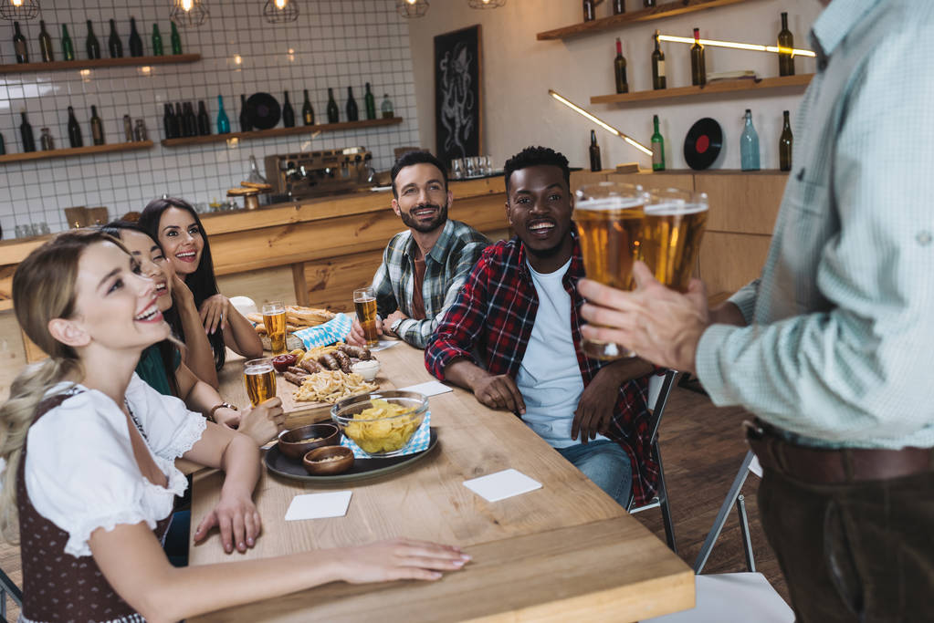 частичный вид человека, держащего бокалы светлого пива возле мультикультурных друзей в пабе
 - Фото, изображение