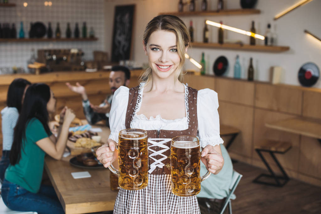 ελκυστική σερβιτόρα σε παραδοσιακό γερμανικό κοστούμι κρατώντας κούπες από ελαφριά μπίρα και χαμογελώντας στην κάμερα - Φωτογραφία, εικόνα