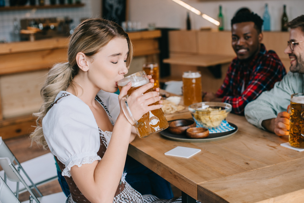 selectieve focus van jonge vrouw in traditioneel Duits kostuum bier drinken in de buurt van multiculturele vrienden - Foto, afbeelding
