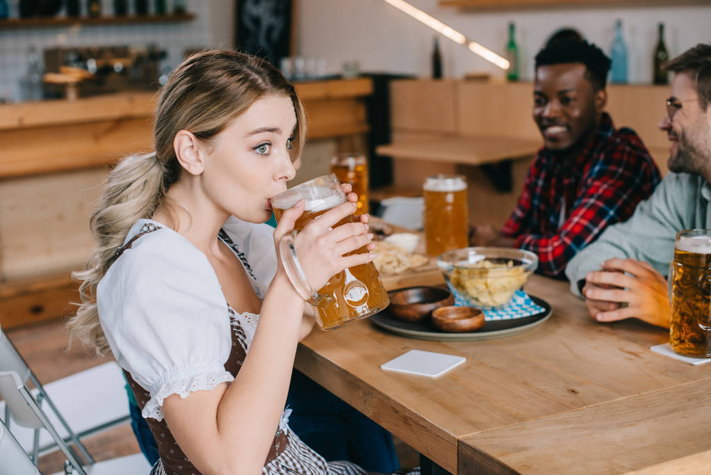 valikoiva painopiste nuori nainen perinteisessä saksalaisessa puvussa oluen juominen lähellä iloinen monikulttuurinen ystäviä
 - Valokuva, kuva