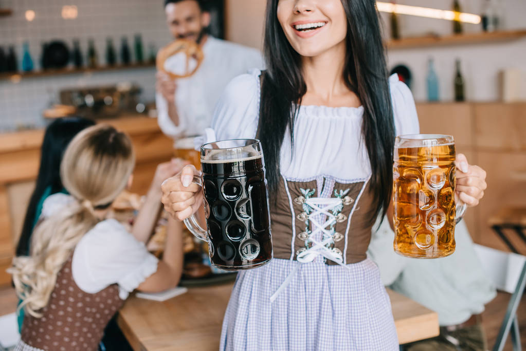 光と暗いビールのマグカップを保持する伝統的なドイツの衣装でウェイトレスのトリミングビュー - 写真・画像