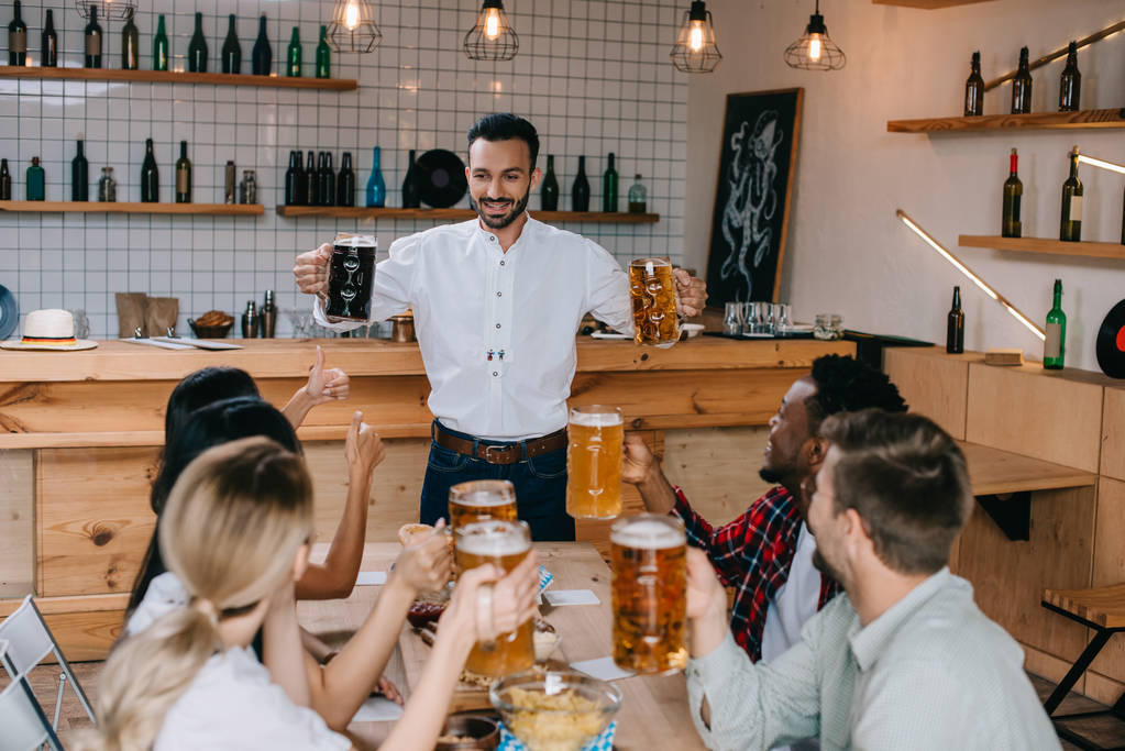 веселий молодий чоловік тримає чашки легкого і темного пива в пабі біля мультикультурних друзів
 - Фото, зображення