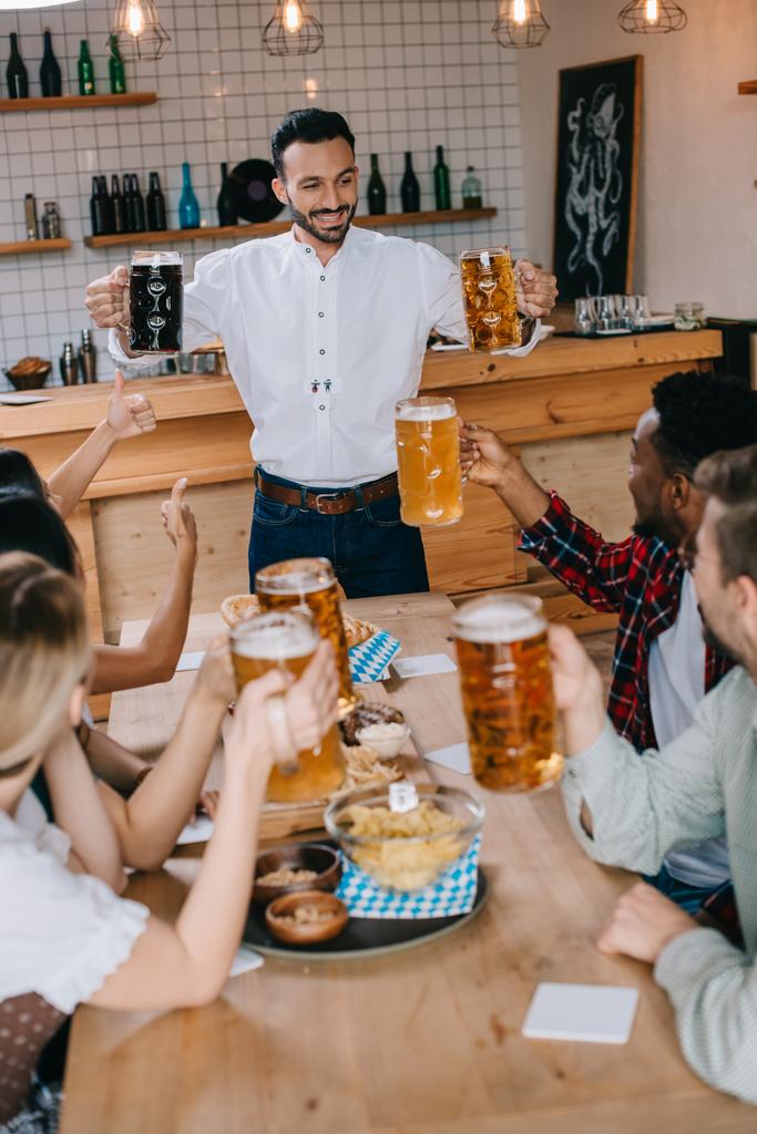 Красивый молодой человек держит кружки светлого и темного пива в пабе рядом с мультикультурными друзьями
 - Фото, изображение