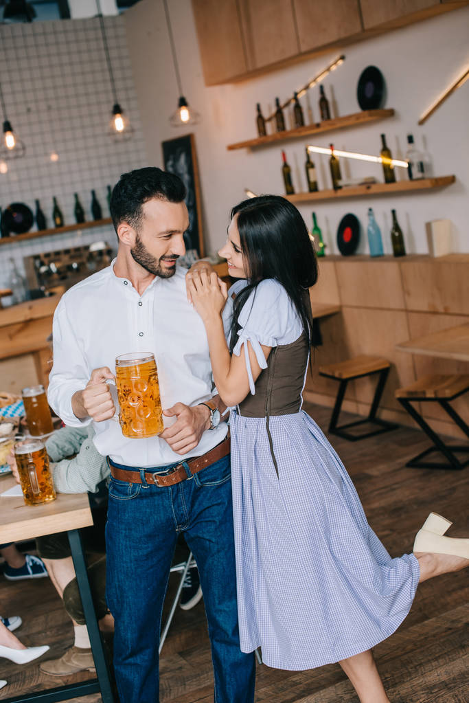 uśmiechnięty młody człowiek trzyma szklankę piwa stojąc w pobliżu pięknej kobiety w tradycyjnym niemieckim stroju - Zdjęcie, obraz