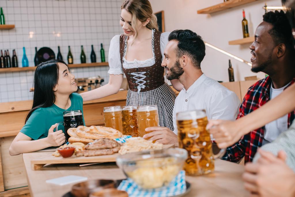 houkutteleva tarjoilija perinteisessä saksalaisessa puvussa, joka tarjoaa olutta monikulttuurisille ystäville pubissa
 - Valokuva, kuva