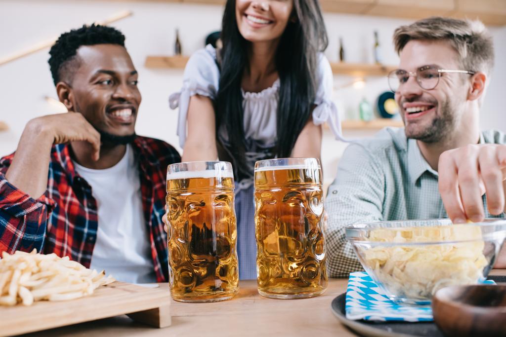 パブで多文化の友人のためにビールを提供する伝統的なドイツの衣装でウェイトレスのトリミングビュー - 写真・画像