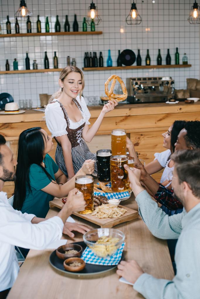 cheerful waitress in traditional german costume holding pretzel while standing near multicultural friends celebrating octoberfest in pub - Photo, Image
