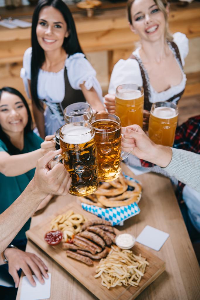 επιλεκτική εστίαση των ελκυστικών σερβιτόρων σε παραδοσιακές γερμανικές στολές που Κουδουνά κούπες μπύρας με πολυπολιτισμικούς φίλους - Φωτογραφία, εικόνα