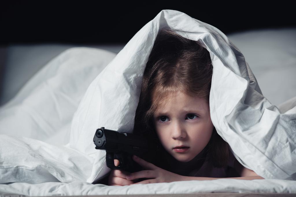 frightened child holding gun while hiding under blanket isolated on black - Photo, Image