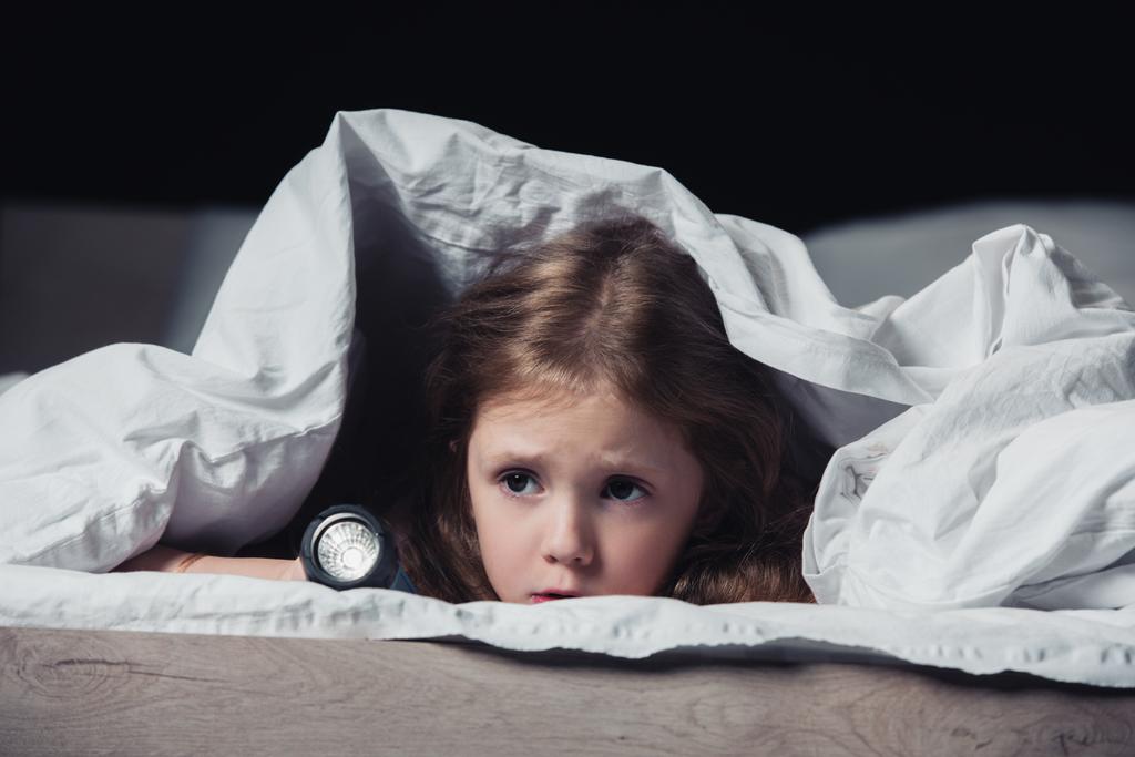 vystrašené dítě ukryté pod přikrývkou a držící svítilnu izolovanou na černém - Fotografie, Obrázek