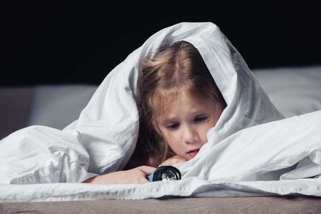 przestraszony dziecko trzyma latarkę ukrywając pod białym kocem na białym tle na czarnym - Zdjęcie, obraz