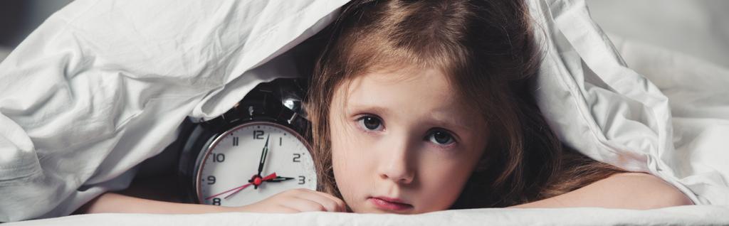 tiro panorâmico de criança assustada escondido sob cobertor com despertador
  - Foto, Imagem