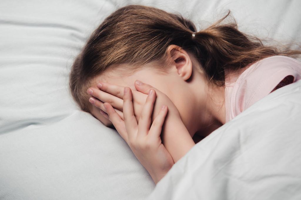 налякана дитина покриває обличчя руками, лежачи на білому ліжку
 - Фото, зображення