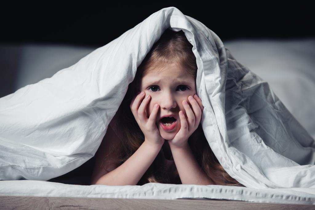 φοβισμένο παιδί ουρλιάζοντας και κρατώντας τα χέρια στο πρόσωπο, ενώ κρύβονται κάτω από την κουβέρτα απομονώνονται σε μαύρο - Φωτογραφία, εικόνα