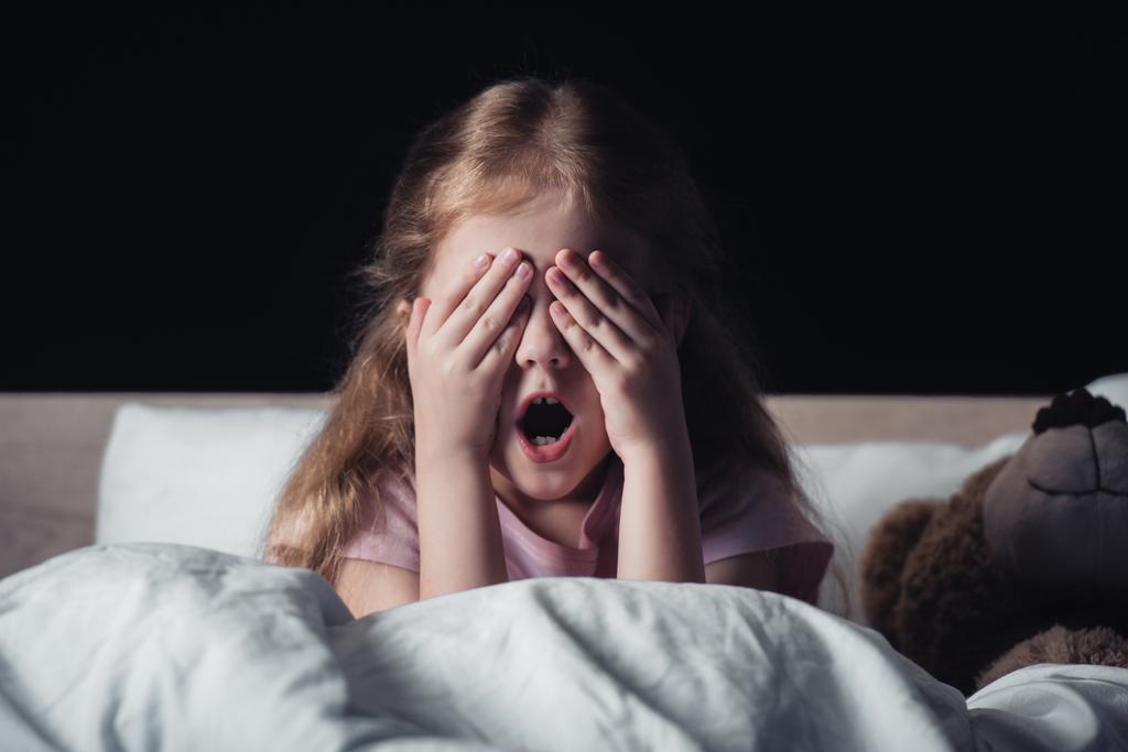 przestraszony dzieciak krzyczeć i pokrycie oczy rękami siedząc na łóżku w pobliżu misia na białym tle na czarno  - Zdjęcie, obraz