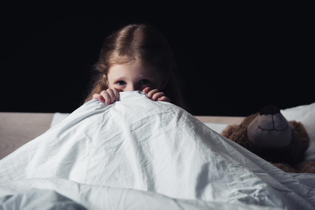 siyah izole oyuncak ayı yakınında yatak üzerinde otururken battaniye nin altında saklanan korkmuş çocuk - Fotoğraf, Görsel