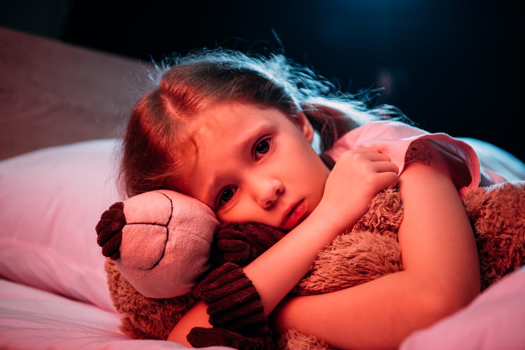 verängstigtes Kind umarmt Teddybär, während es im Bett liegt und isoliert auf schwarz in die Kamera schaut - Foto, Bild