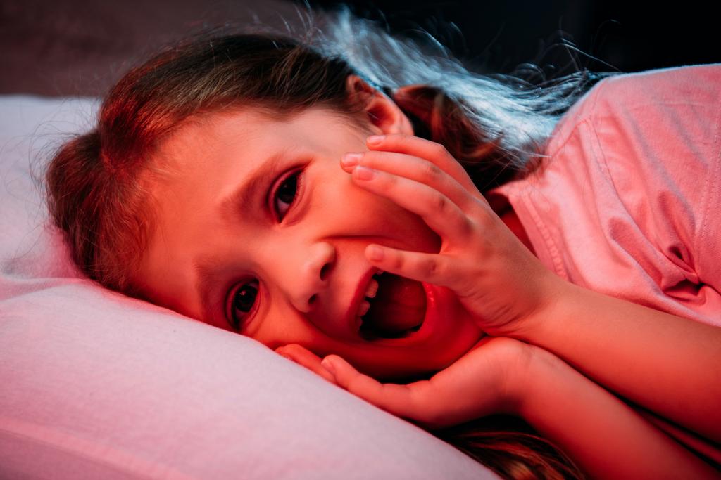frightened child screaming and looking at camera while lying in dark bedroom - Photo, Image