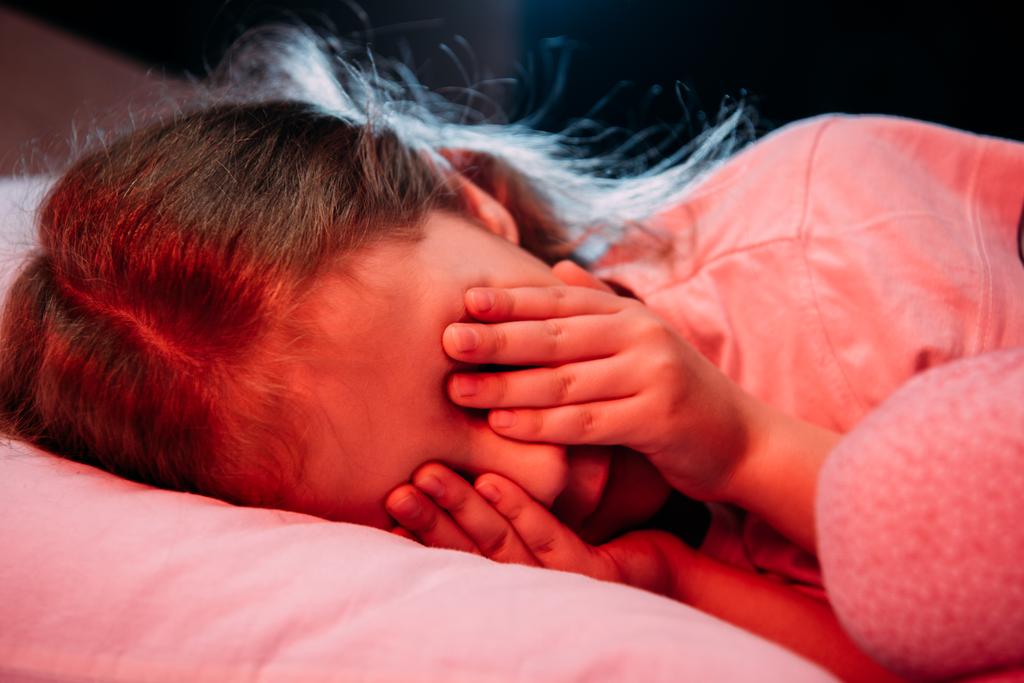 scared child covering eyes with hands while lying in bed isolated on black - Photo, Image