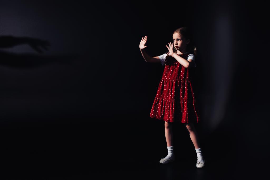 verängstigtes Kind, das mit ausgestreckten Händen in der Dunkelheit vor schwarzem Hintergrund steht - Foto, Bild