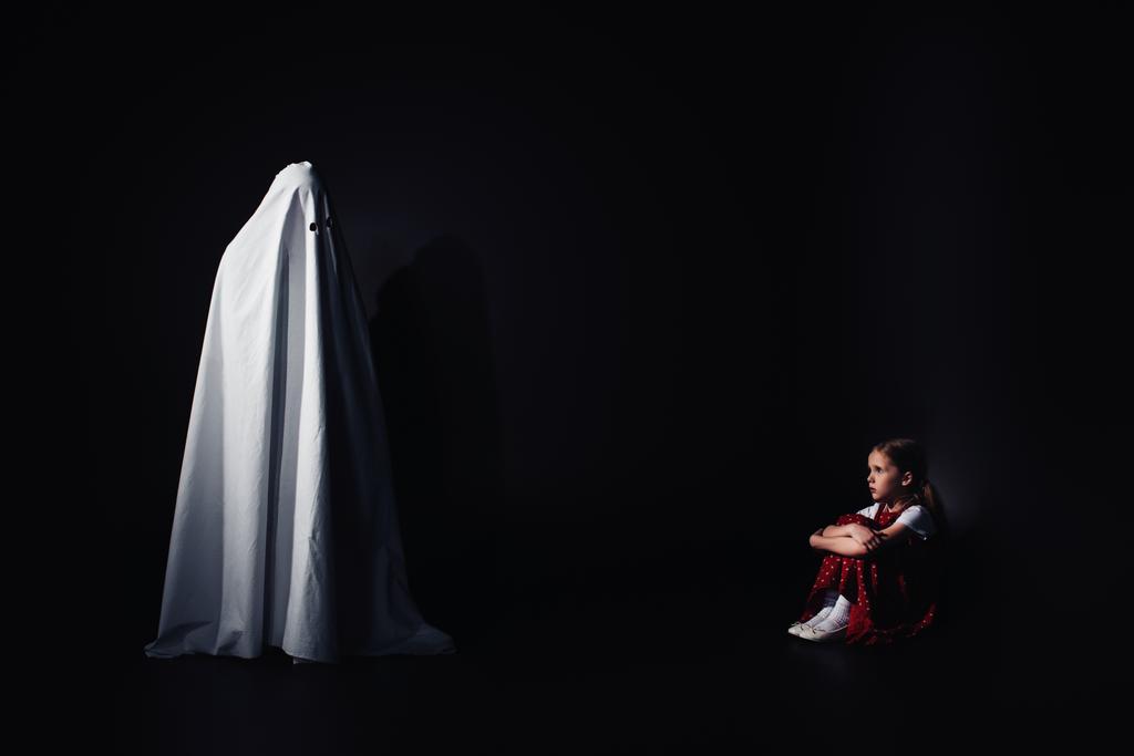 przestraszony dziecko siedzi na podłodze i patrząc na straszny biały duch na czarnym tle - Zdjęcie, obraz