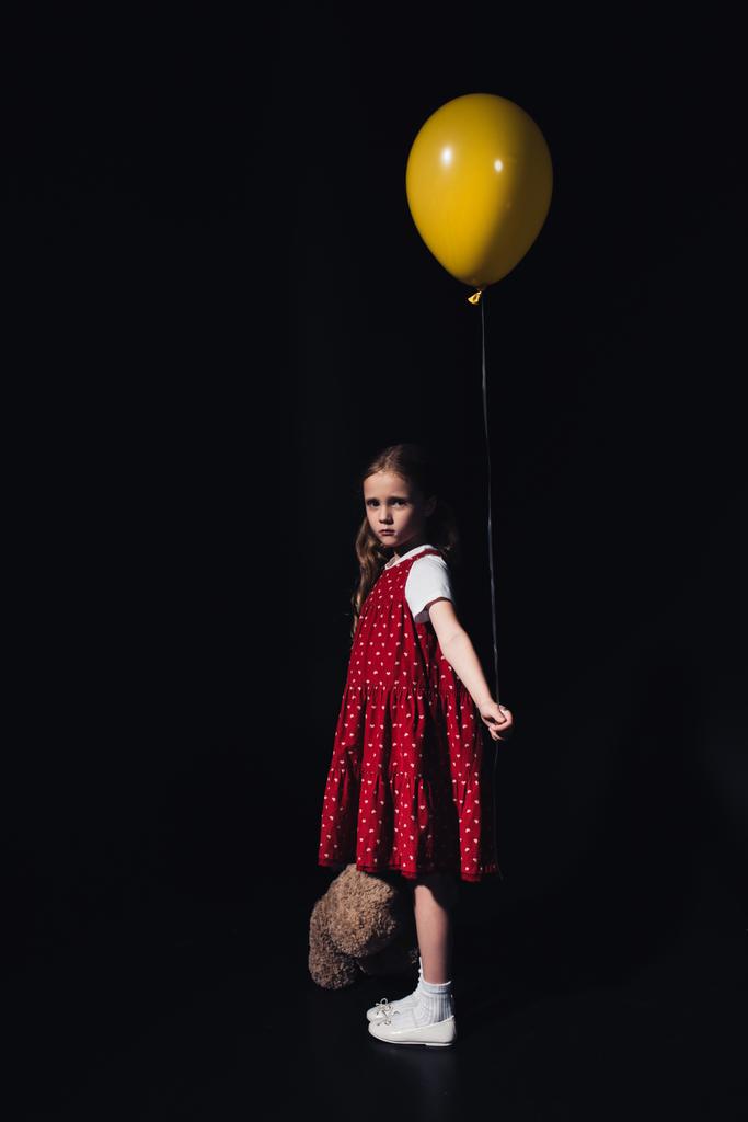 offended child holding yellow balloon and teddy bear while looking at camera isolated on black - Photo, Image