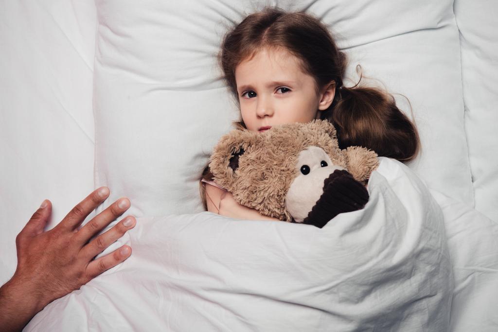 vista recortada de la mano masculina cerca de niño asustado acostado en la cama con oso de peluche
 - Foto, imagen