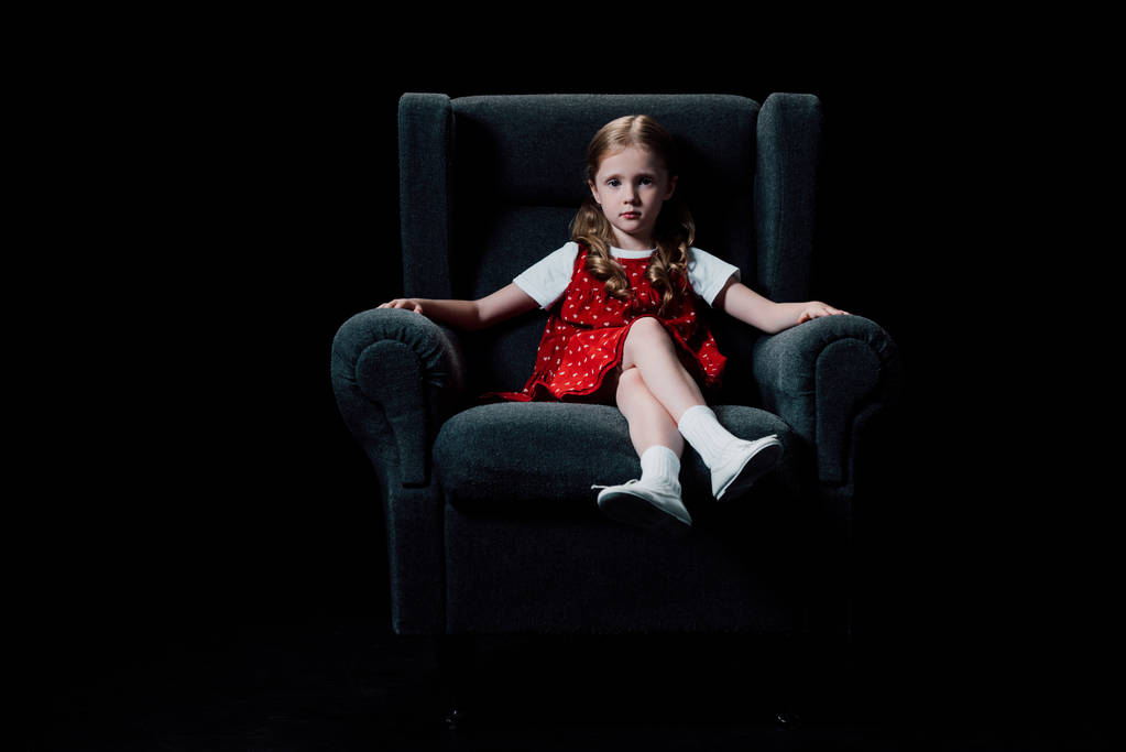 yksinäinen lapsi istuu nojatuolissa ja katselee kameraa eristettynä mustaan
 - Valokuva, kuva