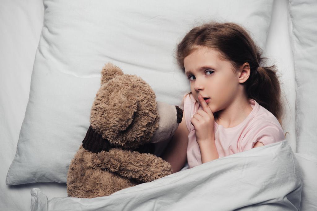 vista superior da criança assustada mostrando sinal de silêncio e olhando para a câmera enquanto deitado na cama com ursinho de pelúcia
 - Foto, Imagem