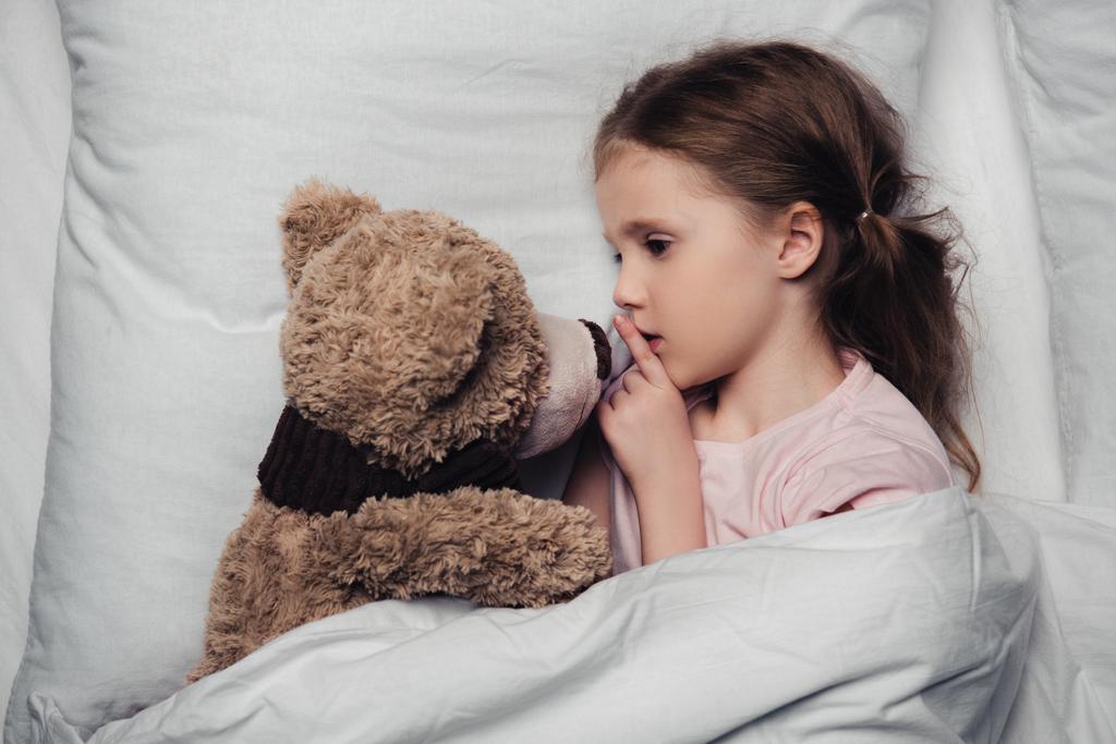 adorable child showing hush sign while lying in bed with teddy bear - Photo, Image