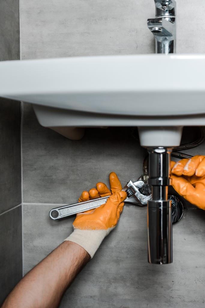 Ausgeschnittene Ansicht eines Reparateurs in Gummihandschuhen, der Wasserschäden mit verstellbarem Schraubenschlüssel behebt - Foto, Bild