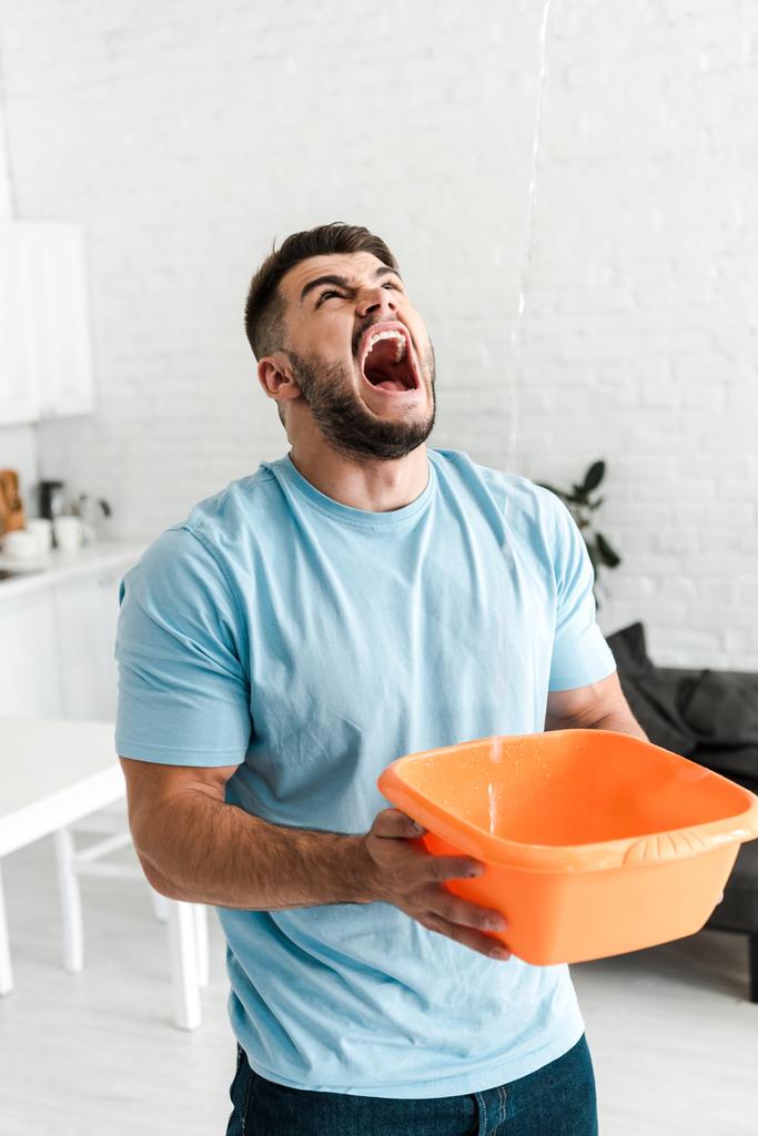 selectieve focus van boos man schreeuwen terwijl het houden van plastic Wash Bowl in de buurt van het gieten van water  - Foto, afbeelding