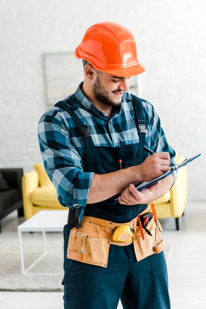 happy bearded worker in safety helmet writing while holding clipboard  - Photo, Image