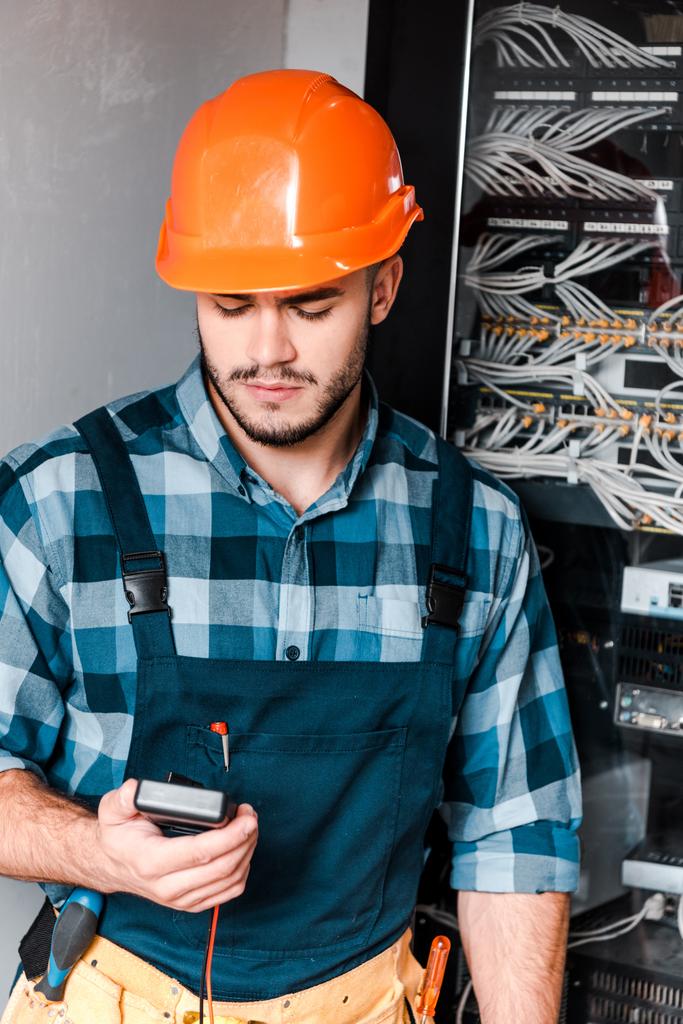 handsome bearded technician holding digital meter near wires and cables  - Photo, Image