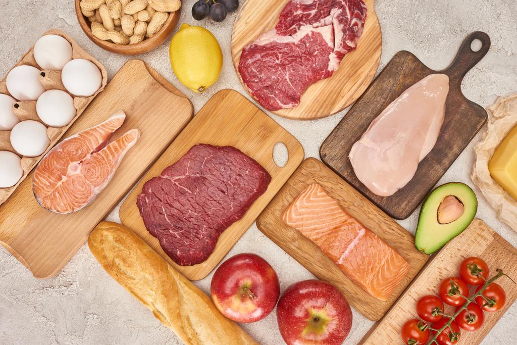 Top view af raw organic assorted meat, poultry, fish, eggs, apples, lemon, avocado, tomatoes and peanuts on wooden cupboards with fresh baguette on marble surface - Photo, Image