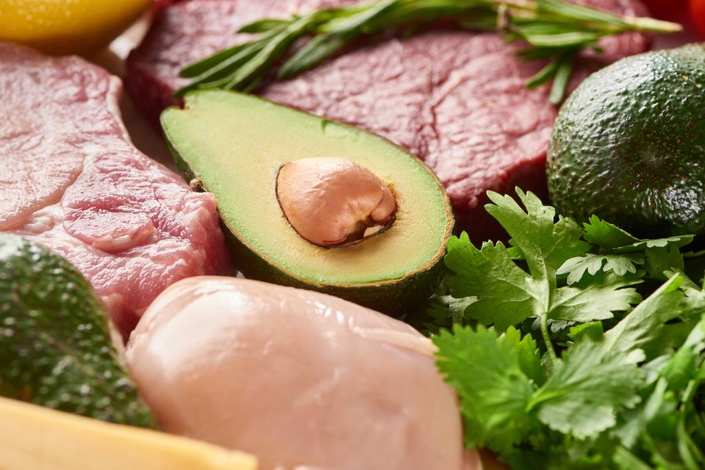 Nahaufnahme der Hälfte der Avocado zwischen rohem Fleisch und Geflügel mit Rosmarin- und Petersilienzweigen - Foto, Bild