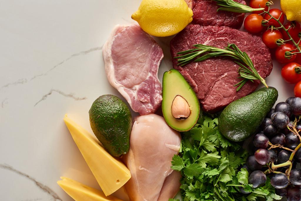 Top view of assorted raw meat and poultry with avocados, lemon, cheese, grapes, branch of tomatoes and greenery twigs - Photo, Image